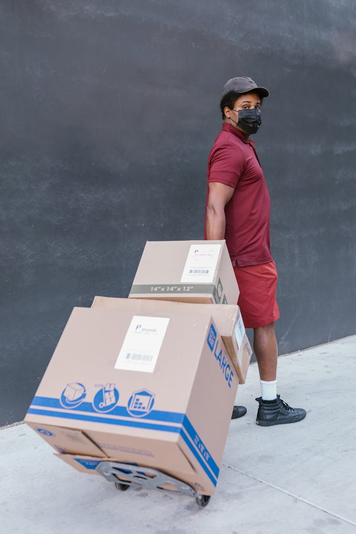 An adult black man wearing a mask pulling a cart with large cardboard boxes outdoors.